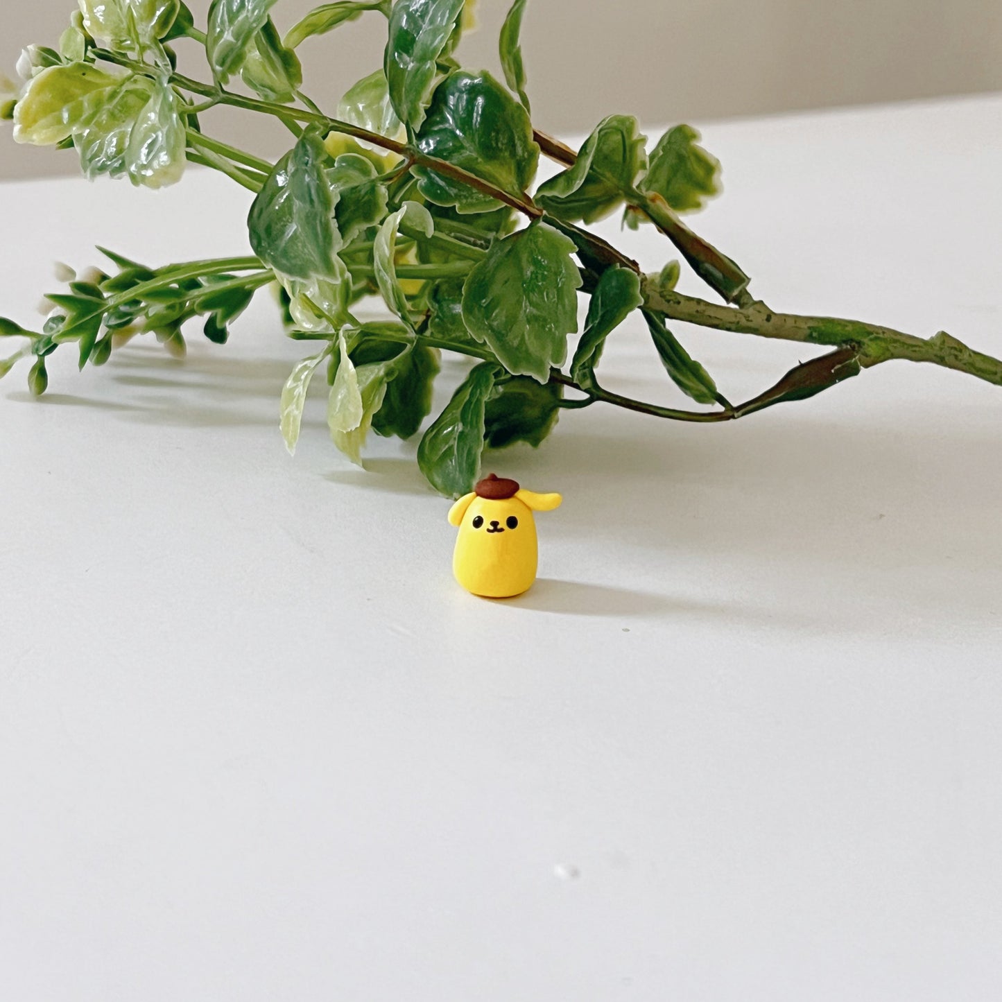 Tiny handmade clay figurine based on Pompompurin. The figurine is sitting on a desk in front of a branch.