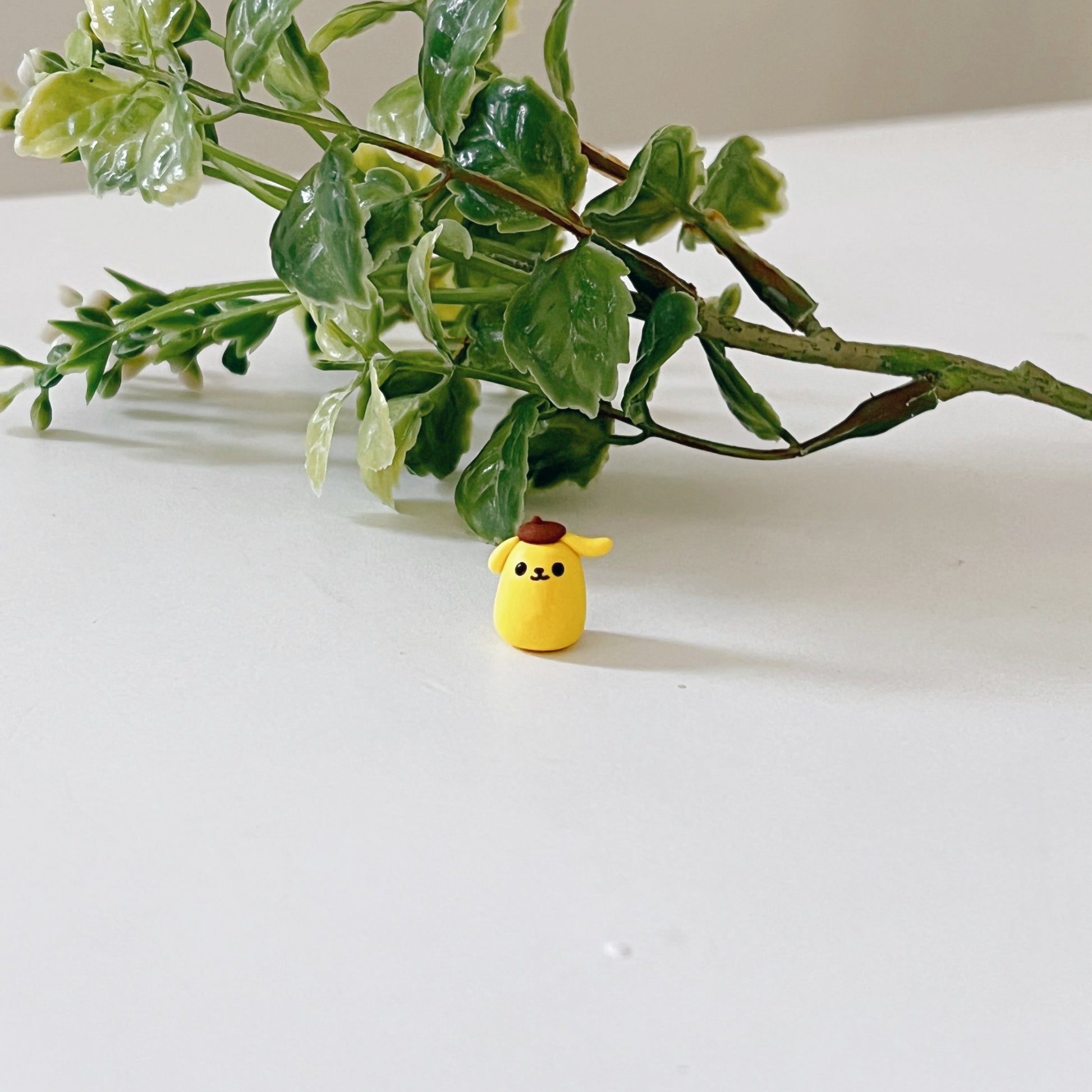 Tiny handmade clay figurine based on Pompompurin. The figurine is sitting on a desk in front of a branch.