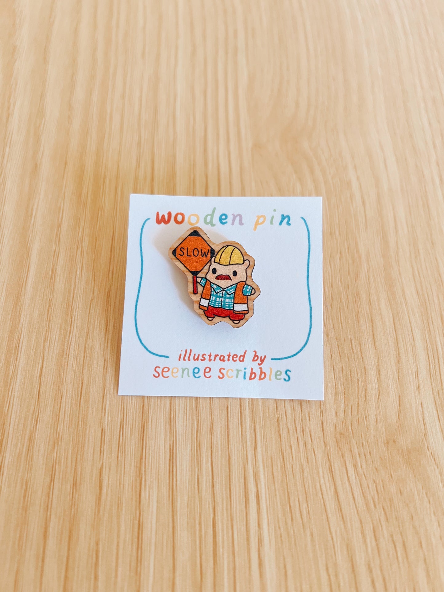 Wooden pin with a construction worker hamster design on a card labeled 'wooden pin' illustrated by Seenee Scribbles, on a wooden surface.