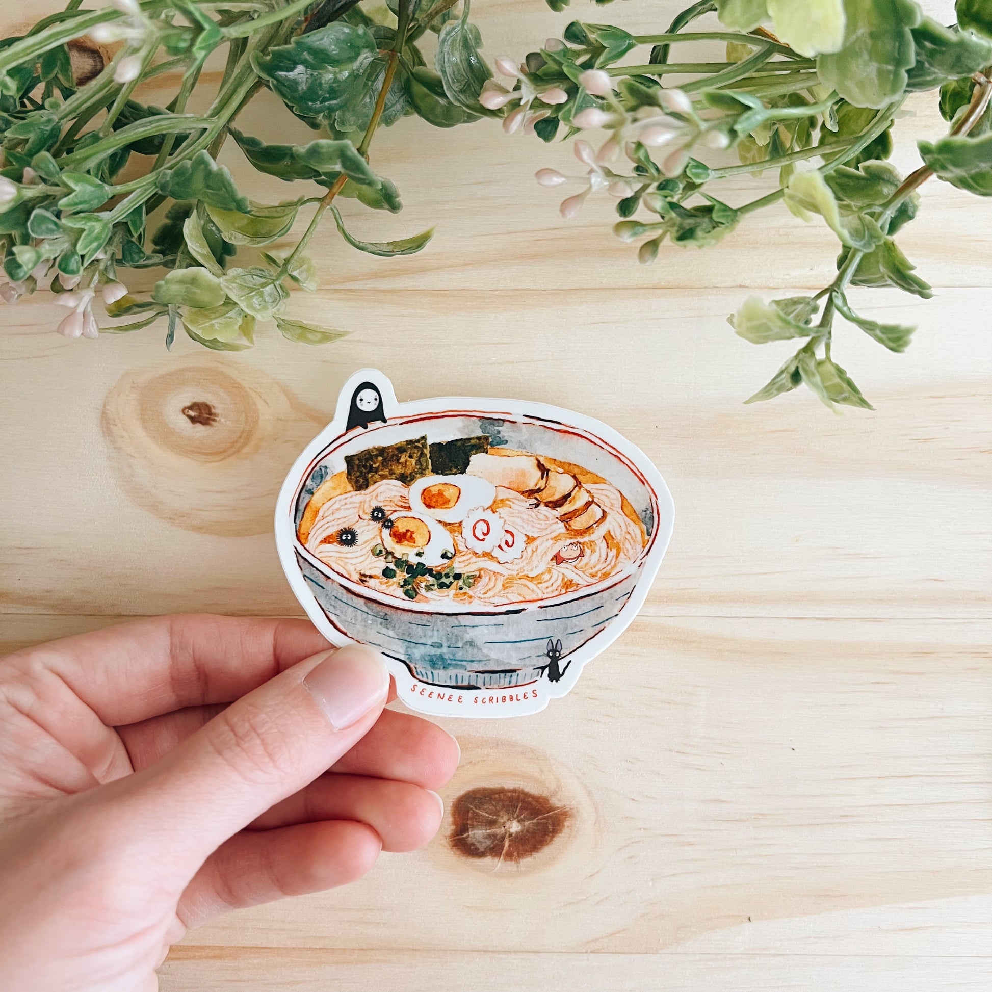 Watercolor painted sticker featuring various characters from Studio Ghibli films. The sticker is being held against a wooden desk with leaves at the top.