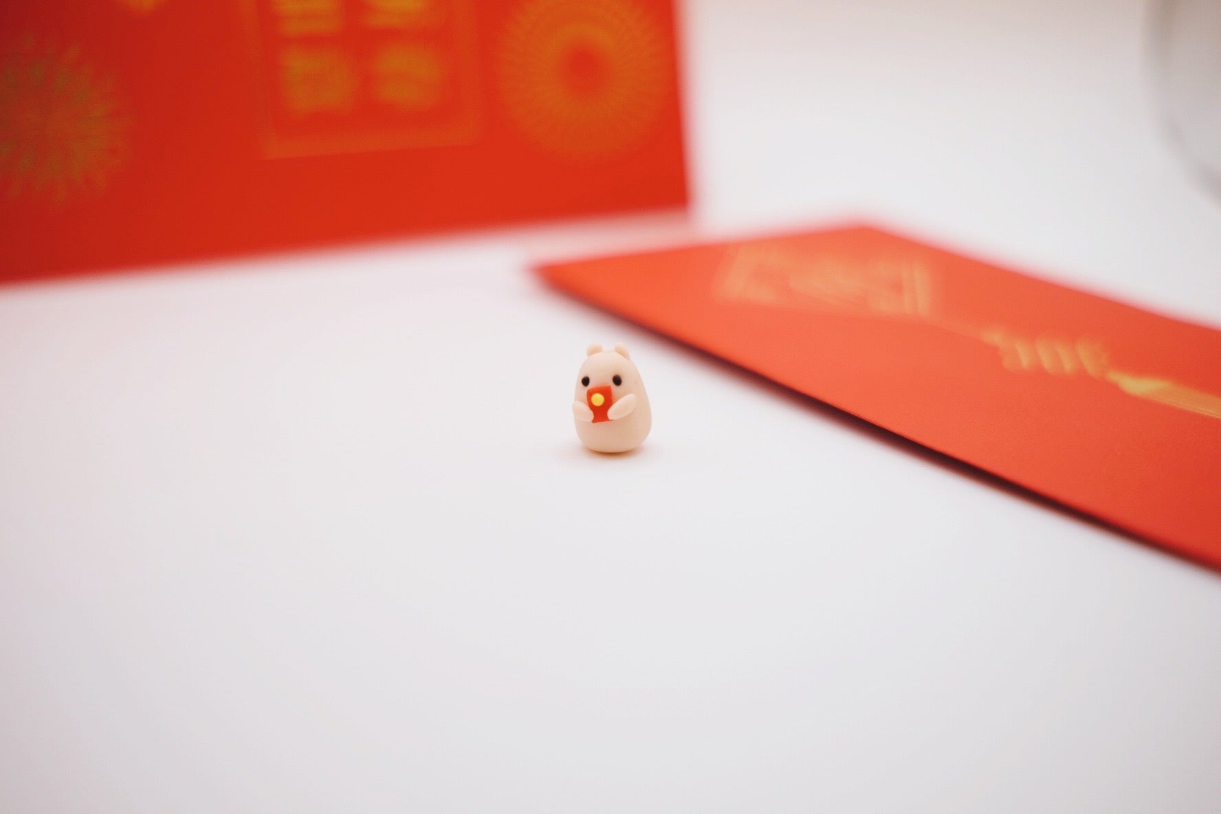 Tiny clay hamster holding a red envelope, with actual red envelopes in the background