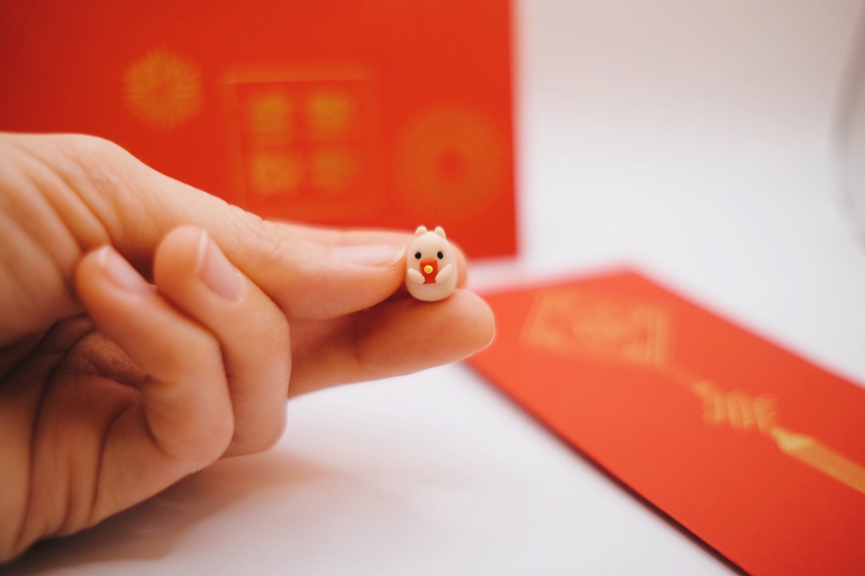 Hand holding a tiny clay hamster with a red envelope. Actual red envelopes in the background.