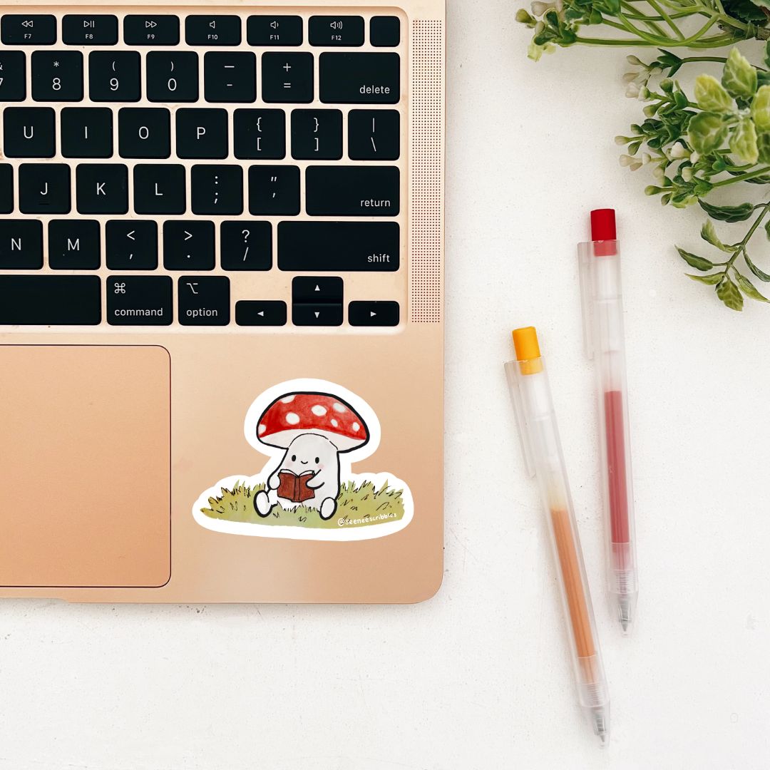 Weatherproof vinyl sticker of a mushroom reading a book, sitting on grass. Hand painted using watercolor and ink. Sticker is depicted on the corner of a laptop on a desk with two pens and leaves.
